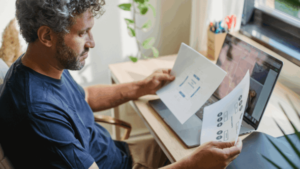 Man reviewing printed charts and diagrams at a desk with a laptop, representing analysis of prior art in the patent process.
