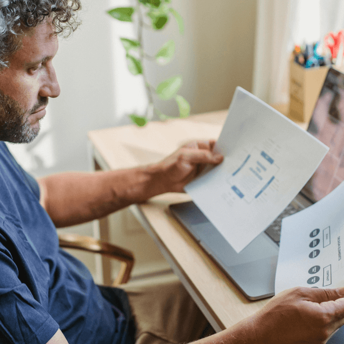 Man reviewing printed charts and diagrams at a desk with a laptop, representing analysis of prior art in the patent process.