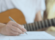 Musician writing in a notebook with a guitar in the background, illustrating the creation of original work.