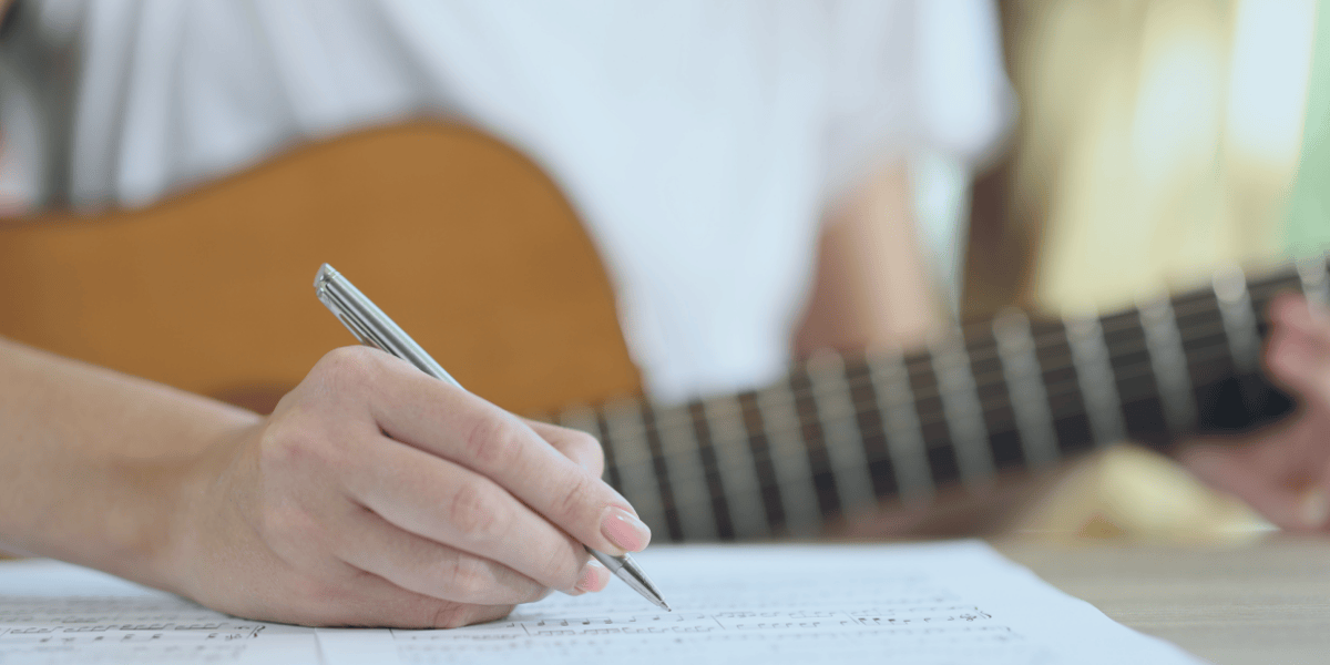 Musician writing in a notebook with a guitar in the background, illustrating the creation of original work.